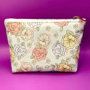 May include: A cream-colored zippered pouch with a floral pattern. The pattern features roses and other flowers in shades of peach, yellow, and white, with green leaves. The pouch has a gold-colored zipper and a cream-colored lining.