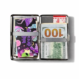 May include: Open silver metal wallet with a clip holding a stack of $100 bills. The left side features a colorful graphic card with the words "high dreams" and an alien illustration. The wallet is open, revealing its contents.