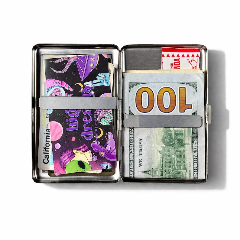 May include: Open silver metal wallet with a clip holding a stack of $100 bills. The left side features a colorful graphic card with the words "high dreams" and an alien illustration. The wallet is open, revealing its contents.