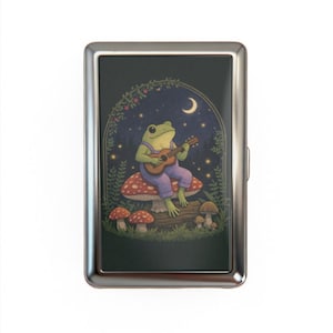 May include: A silver cigarette case featuring a whimsical illustration of a green frog playing a guitar while seated on a mushroom. The background includes a night sky with a crescent moon and stars, and a decorative arch.