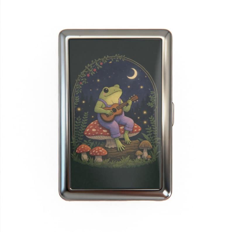 May include: A silver cigarette case featuring a whimsical illustration of a green frog playing a guitar while seated on a mushroom. The background includes a night sky with a crescent moon and stars, and a decorative arch.