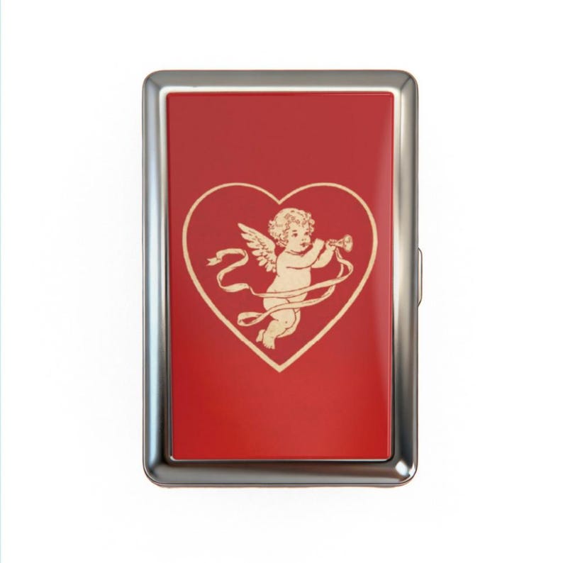 May include: A rectangular, silver-framed cigarette case with a red background. A beige cherub playing a trumpet is inside a heart shape. The case has a silver hinge and clasp.