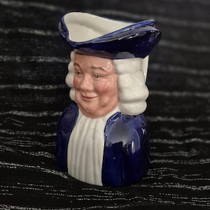 Op de afbeelding: Een keramische karakterkan in de vorm van een persoon met een donkerblauwe driekante hoed en een bijpassende blauwe jas over een wit befje. Het figuur heeft een lichtroze gezicht en witte gepoederde pruikkrullen die de zijkanten omlijsten.