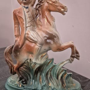 Może przedstawiać: Figurka ceramiczna w stylu vintage przedstawiająca kowboja na koniu. Figurka przedstawia mężczyznę w brązowym płaszczu i kapeluszu, jadącego na brązowym i złotym koniu. Podstawa jest zielona ze złotymi akcentami, a projekt jest szczegółowy i dekoracyjny.