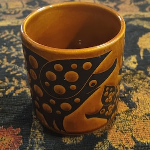 Könnte beinhalten: Ein brauner Keramikbecher mit einem schwarzen, stilisierten Design mit kreisförmigen und organischen Formen. Der Becher hat eine zylindrische Form und eine glänzende Oberfläche. Das Design wiederholt sich um den Becher.