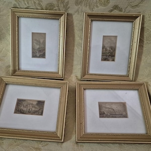 Peut inclure: Quatre cadres dorés contenant des gravures de paysages en noir et blanc. Chaque image est encadrée dans un cadre rectangulaire doré avec un motif légèrement orné, sur fond blanc.