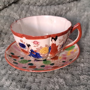 Op de afbeelding: Een vintage theekopje en schotelset met een bloemen- en geisha-ontwerp. Het kopje en de schotel zijn wit met rode, blauwe en gouden accenten. Het ontwerp bevat bloemen, bladeren en twee geisha's in traditionele Japanse kleding.