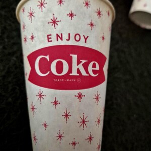 May include: A vintage paper Coke cup with a white background and red snowflake-like starburst patterns. The word "ENJOY" is above a red banner with the word "Coke" in white. The cup has a rolled rim.