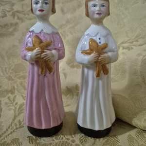 Puede incluir: Dos figuras de cerámica de niñas, una en rosa y otra en blanco, cada una sosteniendo una estrella de mar dorada. Las figuras tienen cabello castaño y visten vestidos con cuellos blancos. Las bases son negras.