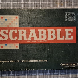 May include: A vintage Scrabble game box with a dark green background. The word "SCRABBLE" is in large white letters on a red rectangle. The game board design is visible on the left. The box includes the text "REGISTERED TRADE MARK".