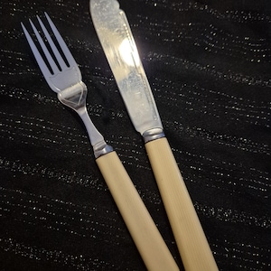 Può includere: Un set di forchetta e coltello da burro con manici color avorio e lame argentate. La forchetta ha quattro rebbi e il coltello ha una punta arrotondata. Il set è esposto su una superficie nera strutturata.