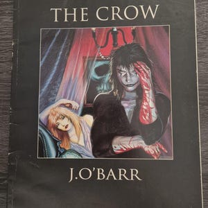 Könnte beinhalten: Ein Taschenbuch mit dem Titel "The Crow" von J. O'Barr. Das Cover zeigt eine dunkle Illustration einer Figur mit Gesichtsbemalung und blutigen Händen, mit einer Frau im Hintergrund. Das Buch ist Band Zwei: Ironie & Verzweiflung.