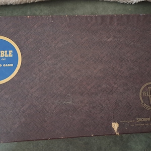 Pode incluir: Uma caixa de cartão castanho com um círculo azul e dourado na frente que diz "SCRABBLE REG. U.S. PAT. OFF. A CROSSWORD GAME". A caixa também tem um círculo dourado com o texto "SEL RIGHT" e as palavras "SELCHOW & RIGHTER CO."