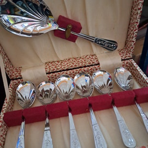 Puede incluir: Un juego de seis cucharaditas de plata con mangos adornados en una caja con estampado rojo y dorado. Las cucharitas están dispuestas en fila, con los mangos hacia arriba. La caja está forrada con una tela blanca.