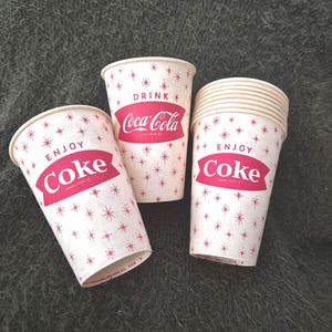 May include: Three vintage Coca-Cola paper cups. The cups are white with a pattern of small red starbursts. The Coca-Cola logo and the words "ENJOY Coke" and "DRINK Coca-Cola" are printed in red.