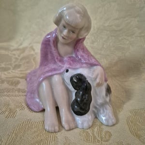Puede incluir: Figura de porcelana de una persona sentada, envuelta en un chal rosa, abrazando a un perro. El perro tiene pelaje blanco y negro. La figura tiene un tono de piel claro y cabello corto y claro. La figura está sobre un fondo amarillo.