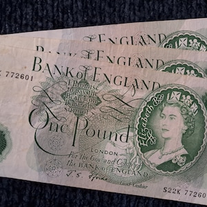 Könnte beinhalten: Ein Stapel von drei Ein-Pfund-Noten der Bank of England mit einem Porträt von Königin Elisabeth II. Die Noten sind grün und weiß mit dem aufgedruckten Text "Bank of England" und "One Pound". Die Seriennummer S22K 772601 ist auf der untersten Note sichtbar.