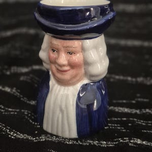 Op de afbeelding: Een keramische karakterkan in de vorm van een persoon met een donkerblauwe hoed en bijpassende jas over een witte bef. Het figuur heeft wit haar, een roze teint en een glanzende afwerking. De bovenkant van de hoed heeft een brede opening.
