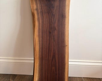 May include: A rectangular wooden serving tray with a live edge and dark brown finish. It features two black metal pipe handles. The tray is displayed against a white wall and brown wood floor.
