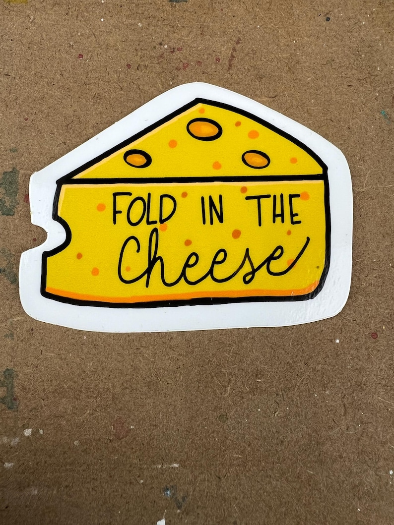 May include: A yellow cheese wedge sticker with black outlines and the text "FOLD IN THE Cheese" in black cursive. The cheese has orange holes and is on a white background.