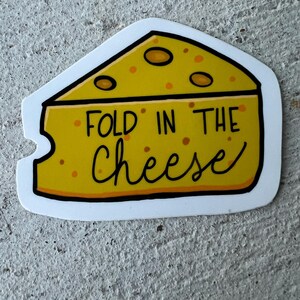 May include: A yellow cheese wedge sticker with the text "FOLD IN THE cheese" in black script. The cheese has orange spots and a white border. The sticker is on a gray, textured surface.