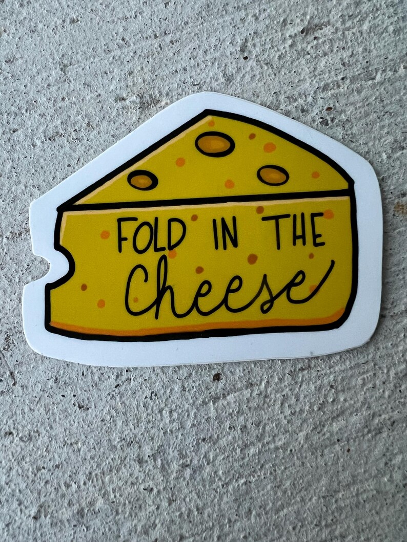 May include: A yellow cheese wedge sticker with the text "FOLD IN THE cheese" in black script. The cheese has orange spots and a white border. The sticker is on a gray, textured surface.