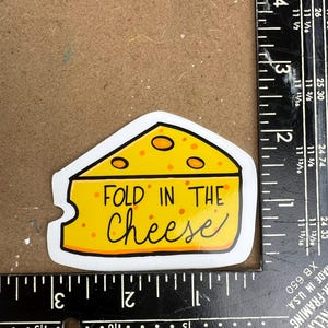May include: A yellow sticker in the shape of a cheese wedge with the words "FOLD IN THE Cheese" in black script. The cheese has orange spots and a white border.