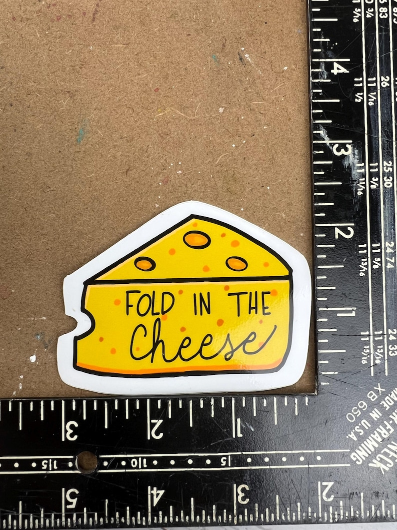 May include: A yellow sticker in the shape of a cheese wedge with the words "FOLD IN THE Cheese" in black script. The cheese has orange spots and a white border.