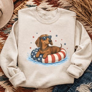 May include: A cream-colored crewneck sweatshirt featuring a graphic of a brown dachshund wearing star-patterned sunglasses. The dog is resting inside a red, white, and blue striped pool float in water, surrounded by small red and blue stars.
