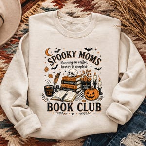 Op de afbeelding: Een crèmekleurig sweatshirt met een grafische print met de tekst "SPOOKY MOMS RUNNING ON COFFEE, HORROR & CHAPTERS BOOK CLUB". Het ontwerp bevat een stapel boeken, een schedel, een pompoen, een koffiemok, kaarsen en vleermuizen.