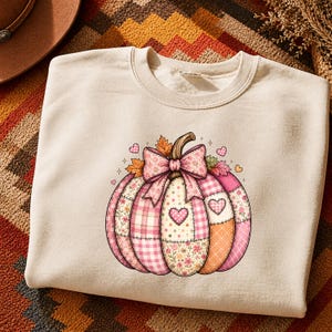 May include: A cream-colored crewneck sweatshirt featuring a central graphic of a patchwork-style pumpkin. The pumpkin is decorated with pink and orange plaid, floral, and polka dot patterns, topped with a large pink bow and small heart accents.