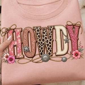 Puede incluir: Una camiseta rosa claro con la palabra "HOWDY" en un diseño de estilo occidental. Las letras tienen acentos florales, de estampado de leopardo y de cuerda, con adornos decorativos y un dije de estrella. Flores rosas y lazos completan el diseño.