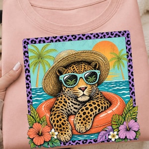 Könnte beinhalten: Hellrosa Sweatshirt mit einem Grafikdruck. Das Design zeigt einen Leoparden mit Sonnenbrille und Strohhut, der in einem orangefarbenen Schwimmring entspannt. Der Hintergrund zeigt eine Strandszene mit Palmen und untergehender Sonne.