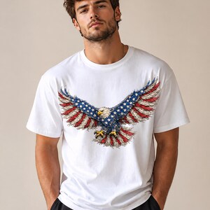 May include: White t-shirt with a detailed graphic of an American bald eagle with outstretched wings. The eagle's wings display the colours of the American flag: red, white, and blue. The shirt is a classic crew neck style.