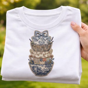 Può includere: T-shirt bianco con un disegno di ciotole decorative impilate. Le ciotole presentano vari motivi, tra cui floreali, paisley e toile. Un fiocco adorna la ciotola superiore. La maglietta è tenuta in mano su uno sfondo verde sfocato.