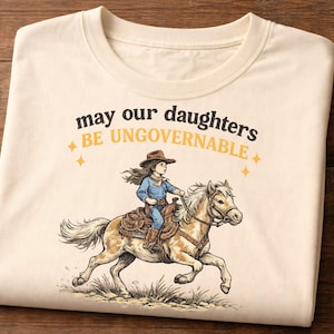 Può includere: T-shirt color crema con grafica di una ragazza a cavallo e la scritta "may our daughters BE UNGOVERNABLE". L'illustrazione utilizza tonalità marroni, blu e beige. La maglietta è piegata su una superficie di legno.