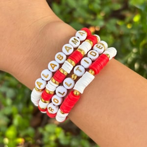 May include: Four beaded bracelets stacked on a wrist. The bracelets are white, red, and gold. The bracelets spell out "ALABAMA ROLL TIDE" with white letter beads. The bracelets are a fan accessory.
