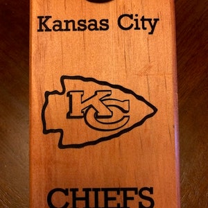 May include: A wooden bottle opener with a black metal top. The opener is engraved with the words "Kansas City Chiefs" and the Chiefs logo.