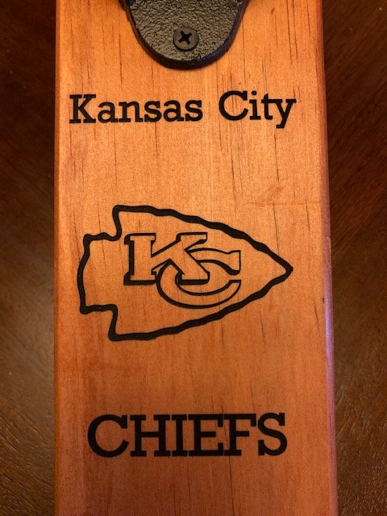 May include: A wooden bottle opener with a black metal top. The opener is engraved with the words "Kansas City Chiefs" and the Chiefs logo.