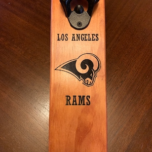 May include: A wooden bottle opener with a black metal bottle opener attached. The bottle opener is decorated with the Los Angeles Rams logo and the words "Los Angeles Rams".