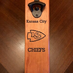 May include: A wooden bottle opener with a black metal bottle opener attached. The bottle opener is engraved with "Kansas City Chiefs".