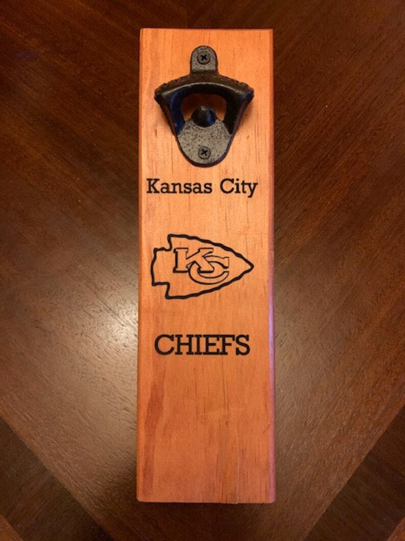May include: A wooden bottle opener with a black metal bottle opener attached. The bottle opener is engraved with "Kansas City Chiefs".