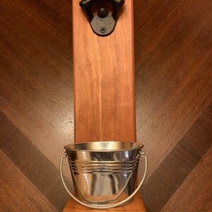 May include: A wooden wall mounted bottle opener with a small metal bucket underneath. The bottle opener is black and the bucket is silver.