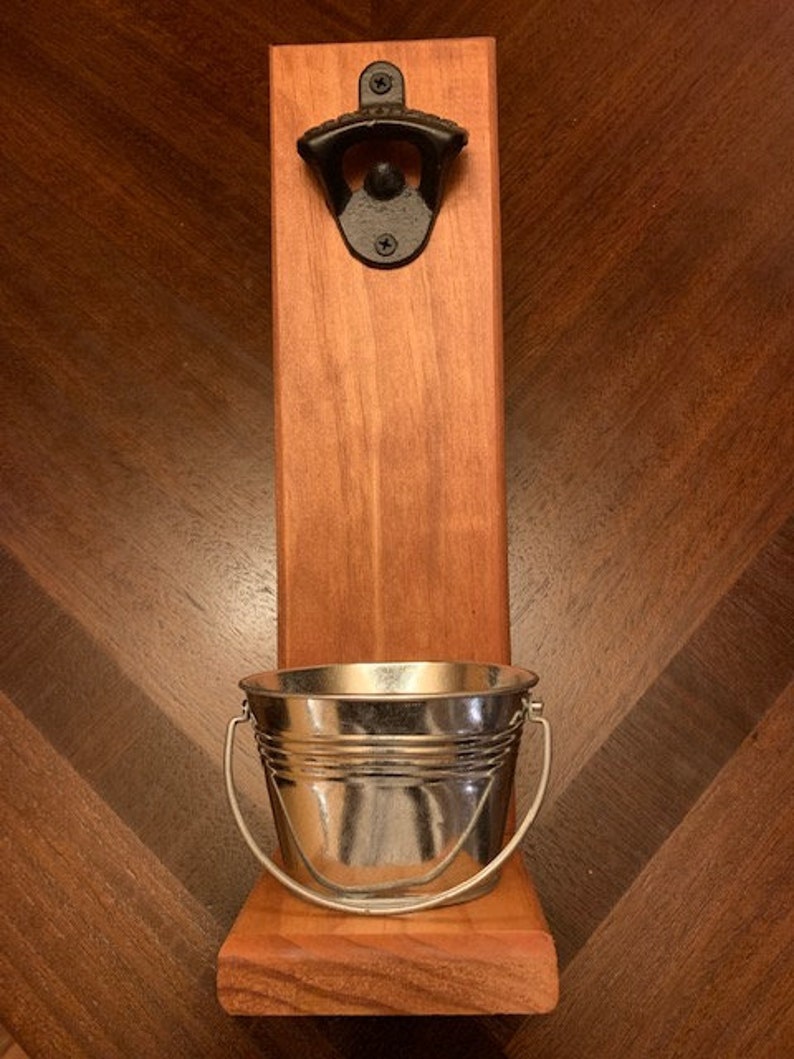 May include: A wooden wall mounted bottle opener with a small metal bucket underneath. The bottle opener is black and the bucket is silver.