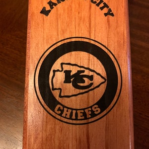 May include: Wooden bottle opener with a black and white Kansas City Chiefs logo. The logo features the team's arrowhead logo and the words "Kansas City Chiefs".