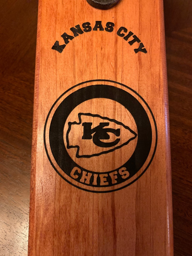 May include: Wooden bottle opener with a black and white Kansas City Chiefs logo. The logo features the team's arrowhead logo and the words "Kansas City Chiefs".