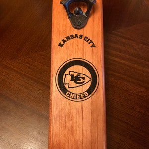 May include: A wooden bottle opener with a black metal bottle opener attached to the top. The bottle opener is engraved with "Kansas City Chiefs" and the Chiefs logo.