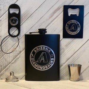 May include: A black flask, bottle opener, and shot glass set. The flask and bottle opener feature the Arizona Diamondbacks logo. The set includes a small metal funnel. The items are displayed on a light wood background.