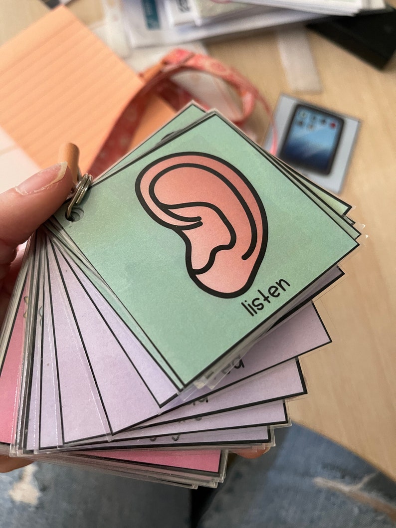 Op de afbeelding: Een stapel flashcards bijeengehouden door een metalen ring. De bovenste kaart toont een roze oorillustratie met het woord "listen" in zwarte tekst op een lichtgroene achtergrond. Andere kaarten zijn zichtbaar.