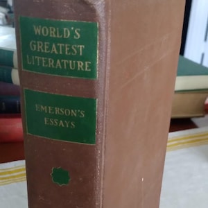 Pode incluir: Um livro antigo de capa dura intitulado "World's Greatest Literature" e "Emerson's Essays". O livro tem uma capa castanha com texto verde e elementos decorativos. A lombada desgastada indica a sua idade.